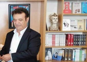 Man in a suit standing near a bookshelf with busts and books.